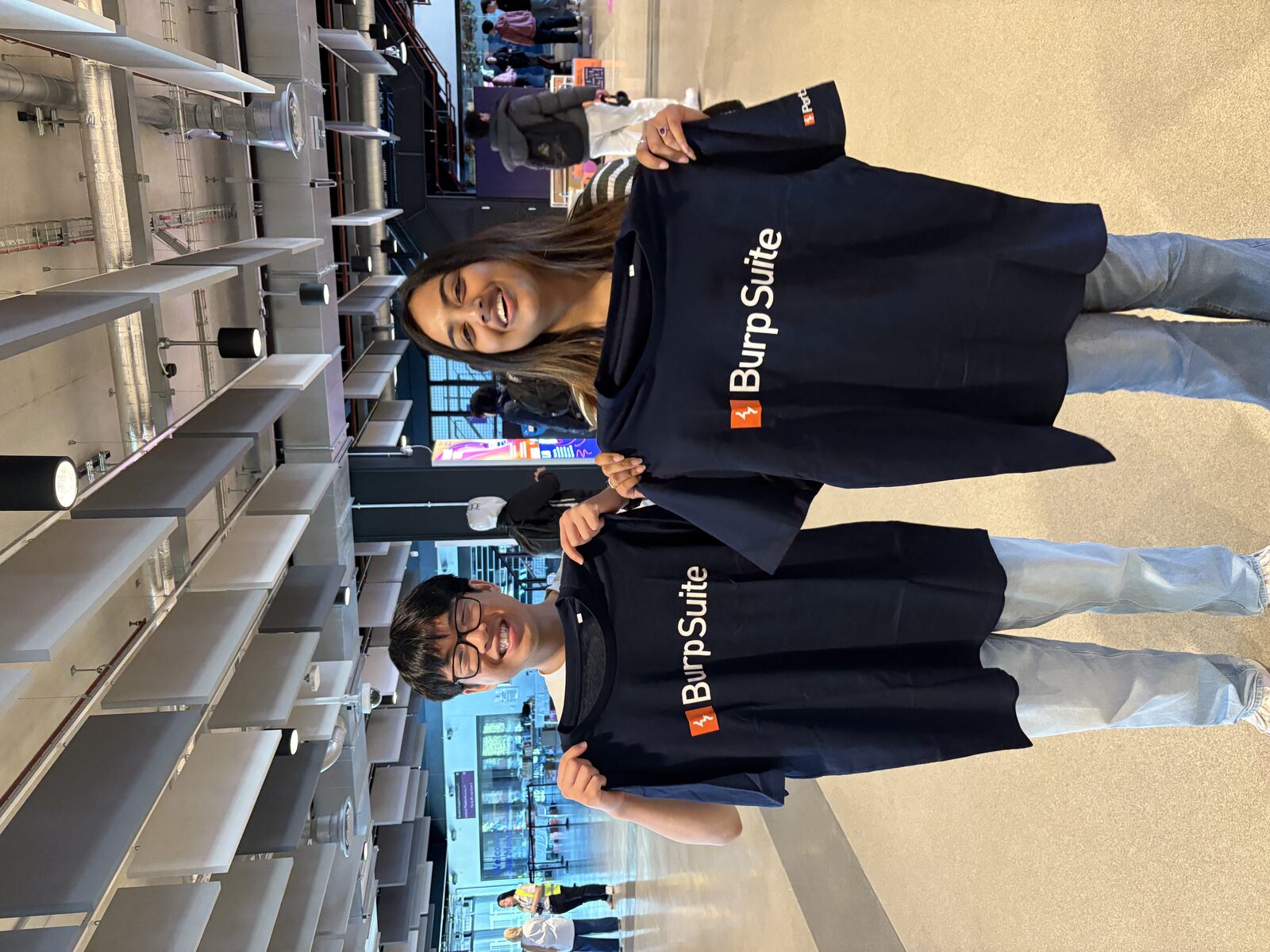 Two students at a hackathon proudly holding up Burp Suite t-shirts
