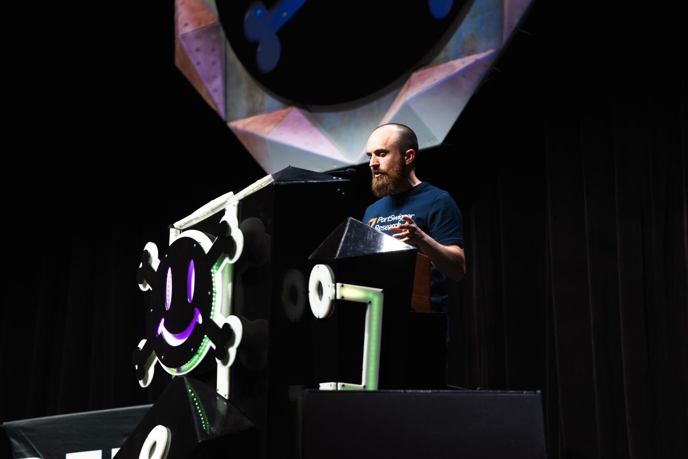 James Kettle on the DEF CON main stage in a PortSwigger Research t-shirt, podium with the DEF CON smiley-skull logo to his left