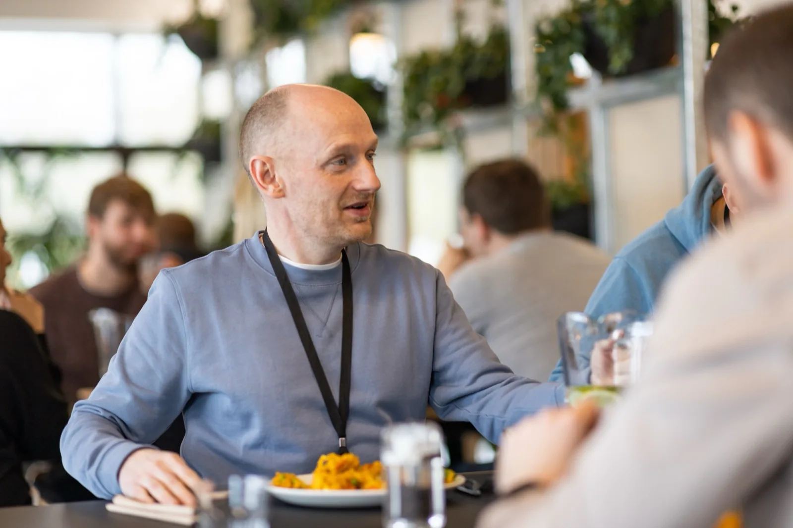 A Swigger mid-conversation at the lunch table, smiling and gesturing, wearing a blue sweater